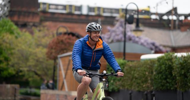 Man wearing helmet and blue jacket smiles as he pedals yellow e-cycle over bridge