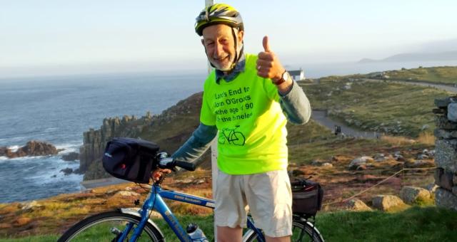 A man stands with a bike under the Land’s End signpost. He is wearing a bright yellow T-shirt and giving a thumbs up to the camera