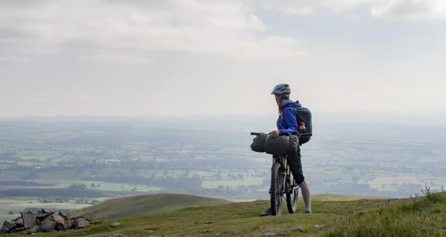 A cyclist straddling her bike. She is looking back at rolling hills behind her. 