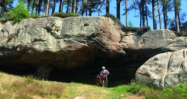 A man is wheeling an orange mountain bike away from a cave. He is wearing cycling kit and a helmet