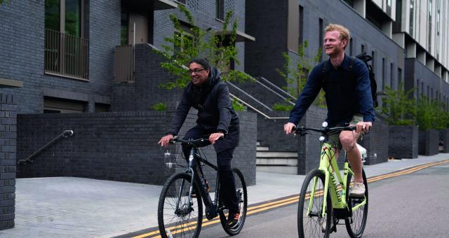 Two men are riding e-cycles side by side along an urban street. They are wearing normal clothes with no cycling helmet.