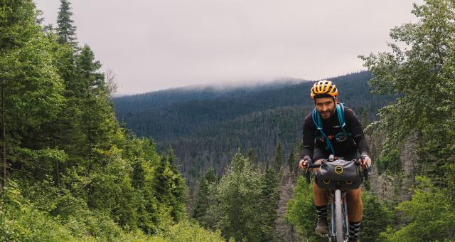 A man is cycling up a gravel path on a gravel bike with a packed handlebar bag. He is wearing black mountain biking kit and a yellow helmet.