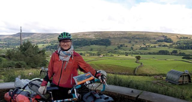 A woman is standing with a fully loaded touring bike. She is wearing a red winter cycling jacket, scarf and green helmet. Behind her is a lot of country scenery