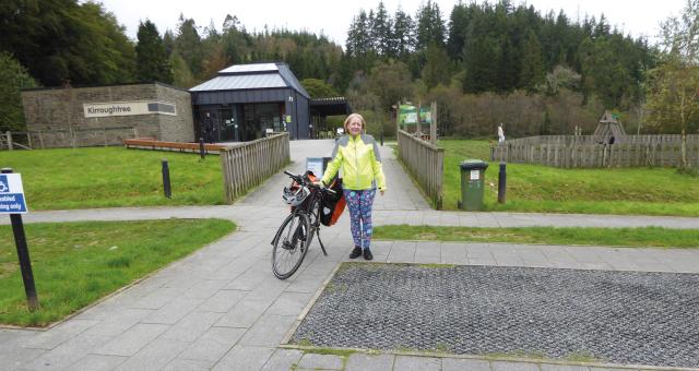 A woman is standing next to her packed touring bike in a park. She is wearing patterned leggings and a hi-vis yellow jacket.