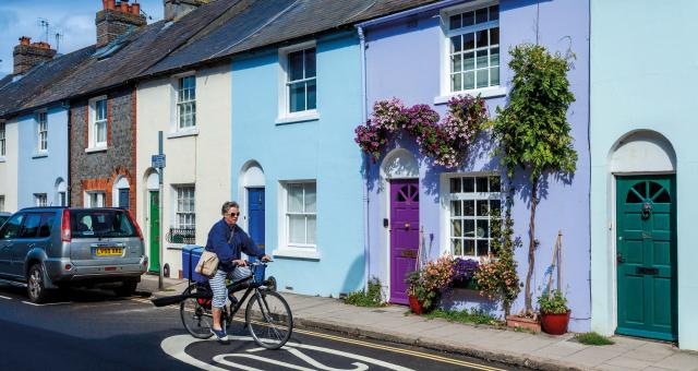 A woman wearing blue and white striped three-quarter length trousers and a blue top is cycling over a 20 MPH logo painted on a road. She is going past a row of pastel painted houses. She is wearing sunglasses and has a shoulder bag across her body