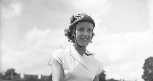 Eileen Sheridan is pictured in a black and white photo at Herne Hill cycle track in London. She is sitting on her Hercules bike wearing shorts and a short-sleeved top and cycle helmet and is smiling