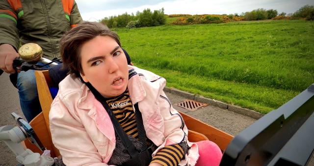 Greta Rafi McMillan is wearing a pink jacket sitting on the front of a cargo bike. There is a grassy hill in the background