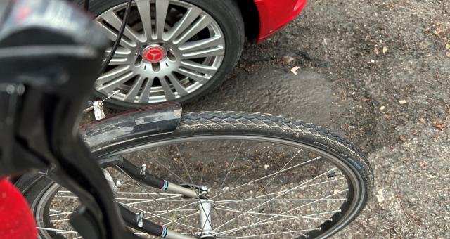 A large pothole in an urban street with a red car's wheel in it and a black bike wheel next to it