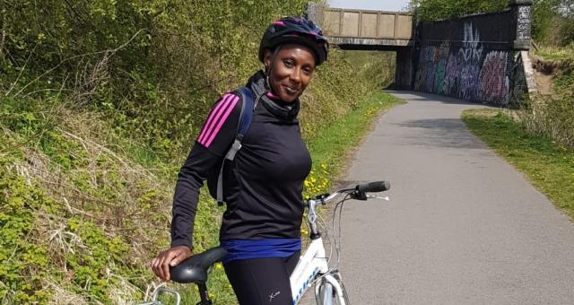 A woman is standing astride a hybrid biked on a paved path. She is wearing cycling kit and helmet