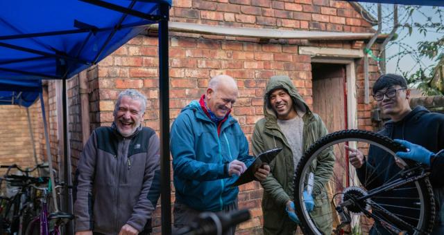 Four men are seen standing under a marquee. They are laughing amongst themselves and smiling at the camera, one holds a bicycle wheel and another holds a clipboard