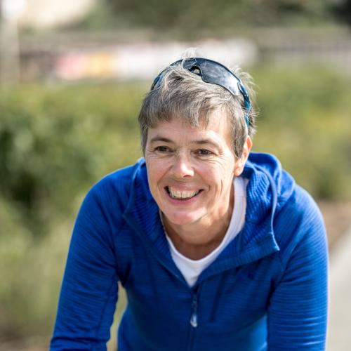 A woman is sitting on her bike with her forearms resting on the drop handlebars, her hands clasped in front of her. She is wearing a blue fleece and has sunglasses on her head. She has short hair. She is smiling.