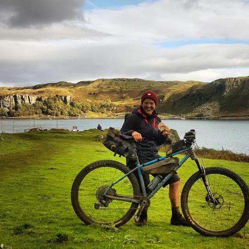 A woman is standing with a very muddy bike and holding a medal, having just completed an off-road triathlon on the Isle of Kerrera. She is smiling broadly